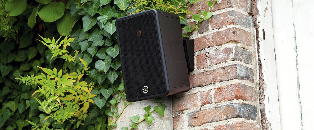 Musta Monitor Audio Climate 50 -ulkokaiutin asennettuna tiiliseinään vihreän kasvillisuuden ympäröimänä