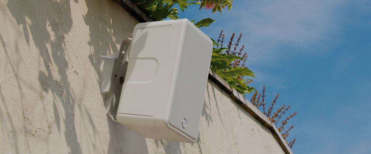 Valkoinen Monitor Audio Climate 60 -kaiutin kiinnitettynä ulkoseinään, sinistä taivasta ja kasveja taustalla – IP55-säänkestävä ulkokaiutin auringonvalossa